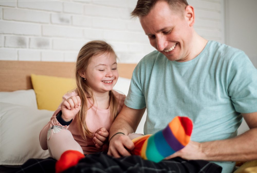 Mismatched socks align with World Down Syndrome Day; pictured: smiling dad puts mismatched socks on his smiling daughter, who has Down syndrome